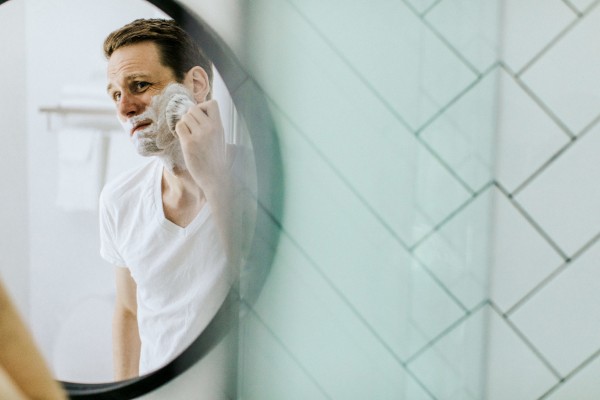 man shaving his beard in mirror of bathroom with white subway tile
